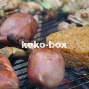 キャンプ場でバーベキュー。骨付き肉と焼きおにぎり、焼き鳥を炭火で焼いている写真