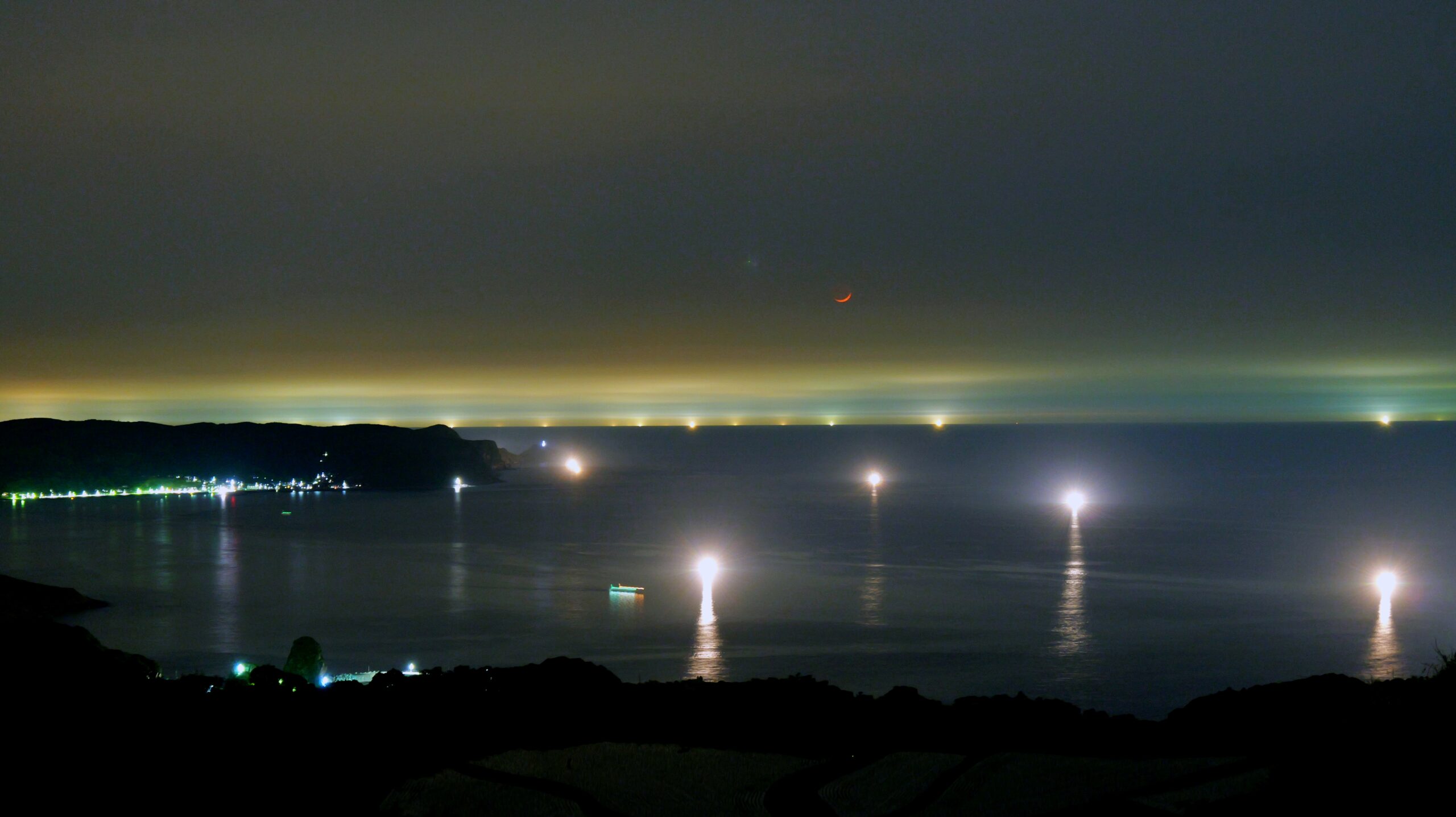新月の日に赤く光る月と漁火と街の光がきれいな写真