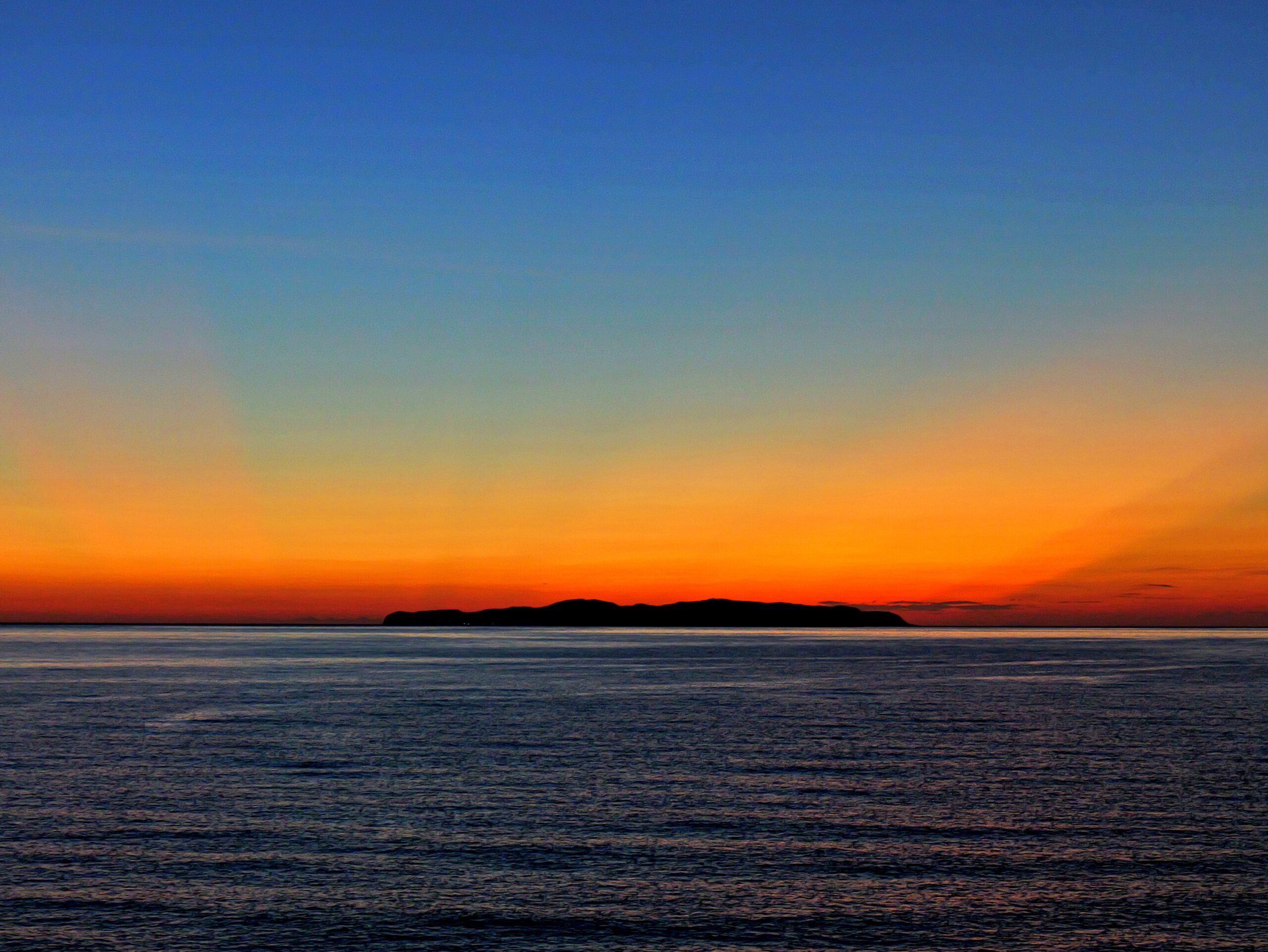 水平線と島から伸びる赤とオレンジの射線が綺麗なグラデーションの夕焼けの写真