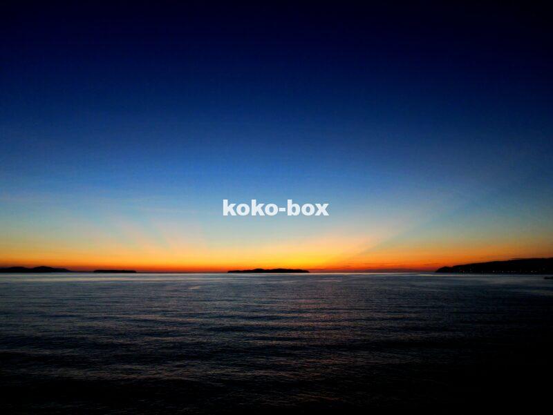 水平線と島から伸びる赤とオレンジの幻想的な射線が綺麗なグラデーションの夕焼けの写真