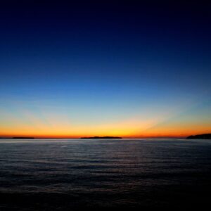 水平線と島から伸びる赤とオレンジの幻想的な射線が綺麗なグラデーションの夕焼けの写真