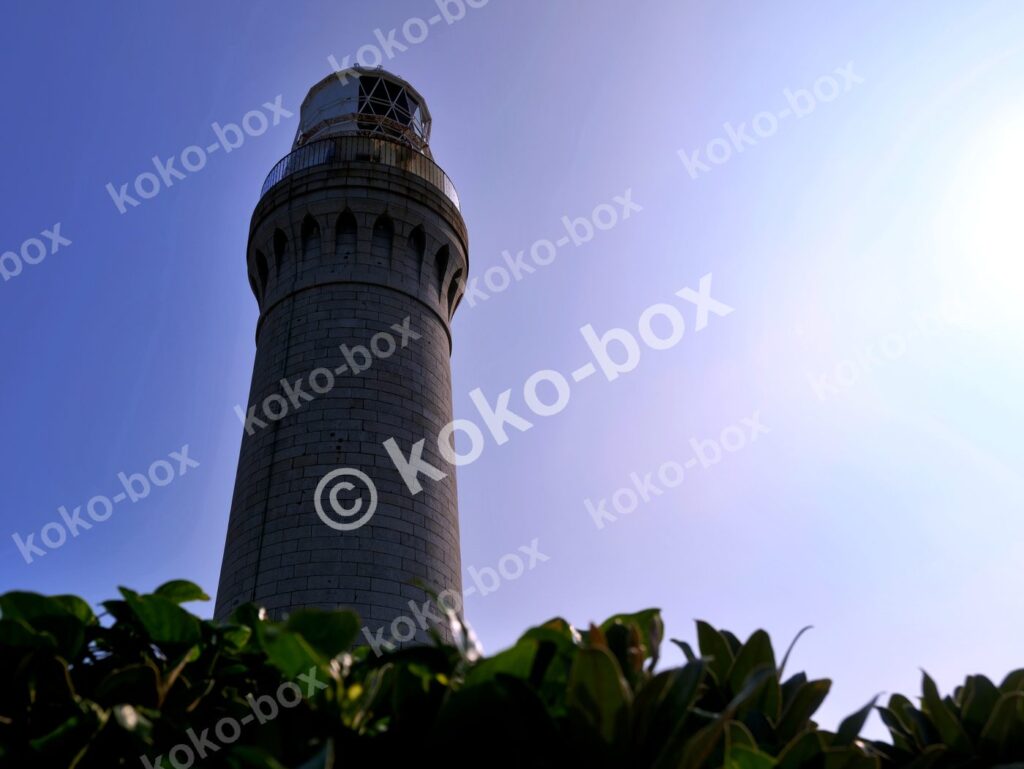 青空を背景に建つ石造りの灯台と前景の緑（横位置）