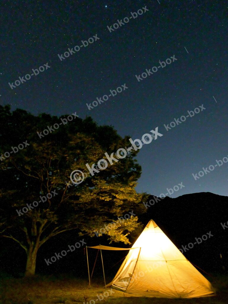 満天の星空の下で温かく光るワンポールテントと大きな木の縦位置写真